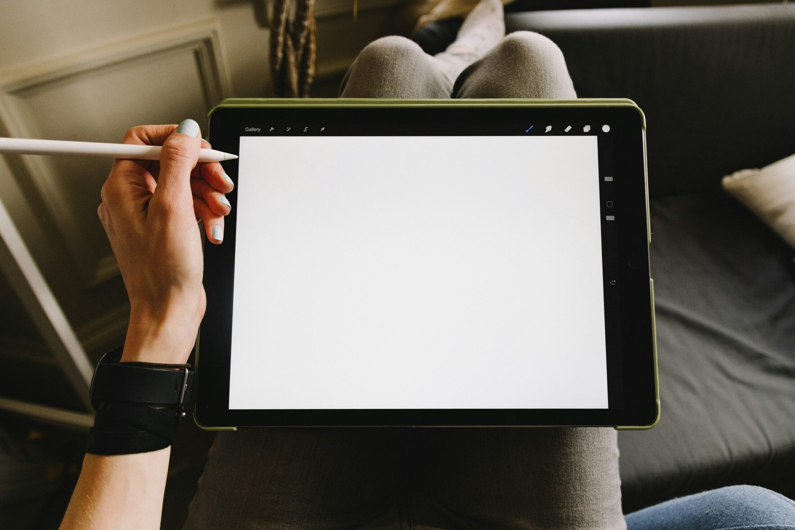 Osoba trzymająca cyfrowy tablet z pustym ekranem i stylusem w dłoni, siedząca z nogami skrzyżowanymi na kanapie. Aplikacja do rysowania jest uruchomiona na tym urządzeniu.