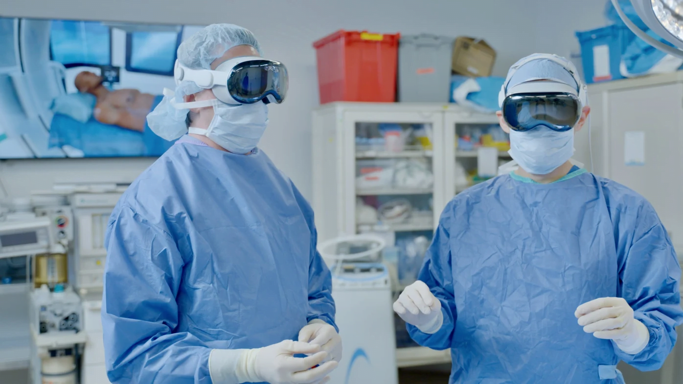 Dwóch lekarzy w niebieskich fartuchach operacyjnych i goglach Apple Vision Pro w sali operacyjnej, w tle wyświetlany jest obraz z sali operacyjnej.