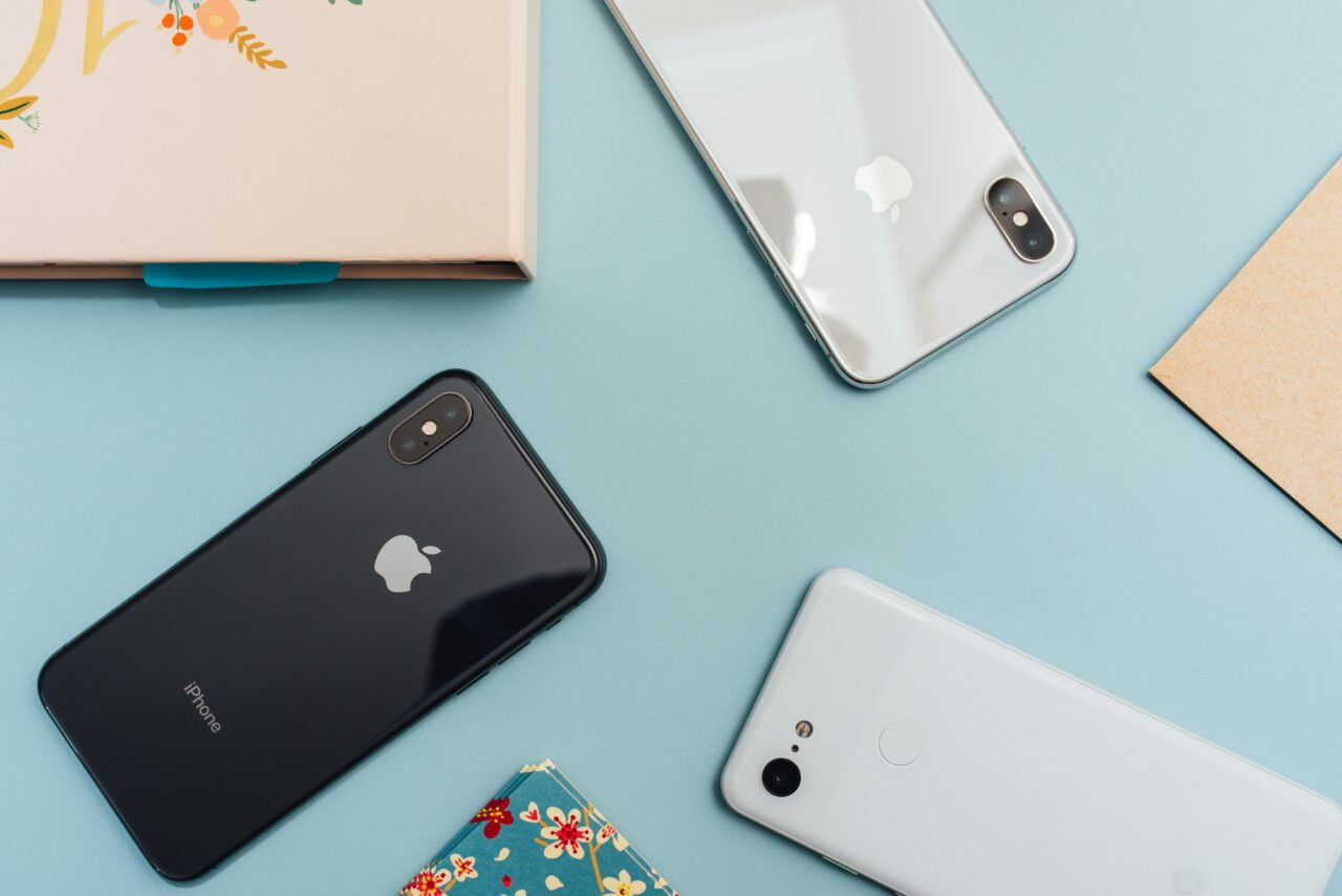 Trzy smartfony Apple leżące na błękitnym tle obok notatnika i kawałka kartonu.
