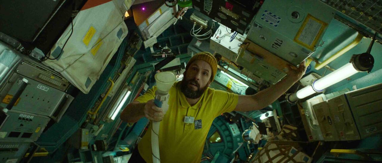 Zdjęcie z filmu Astronauta od Netflix. Mężczyzna w żółtej koszulce i żółtej czapce pływa w symulowanym mikrograwitacyjnym środowisku przypominającym wnętrze stacji kosmicznej, trzymając biały przedmiot przypominający megafon.