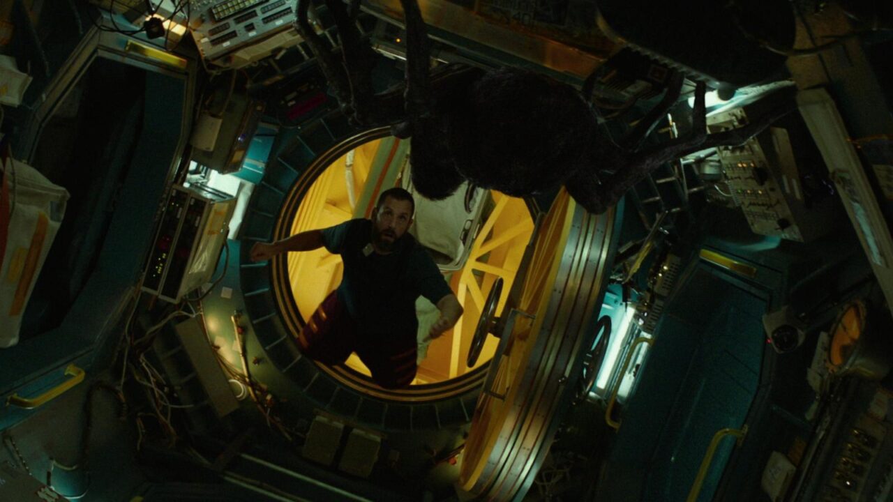 Zdjęcie z filmu Astronauta od Netflix. Mężczyzna w stacji kosmicznej stoi w luklu przy otwartych, żółtych drzwiach, za nim unosi się duża, czarna istota przypominająca pająka. Pomieszczenie wyposażone jest w panele kontrolne i sprzęt techniczny.