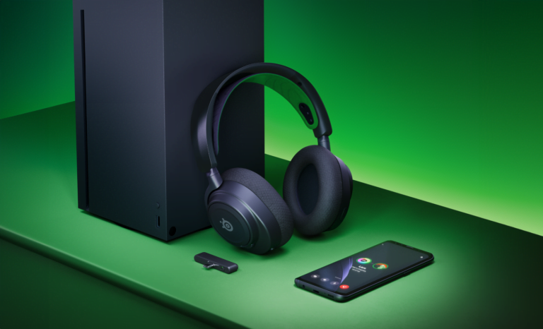 Czarny zestaw gamingowy na zielonym tle, w tym konsola gier wideo, bezprzewodowe słuchawki, pendrive i smartfon z widocznym ekranem początkowym.