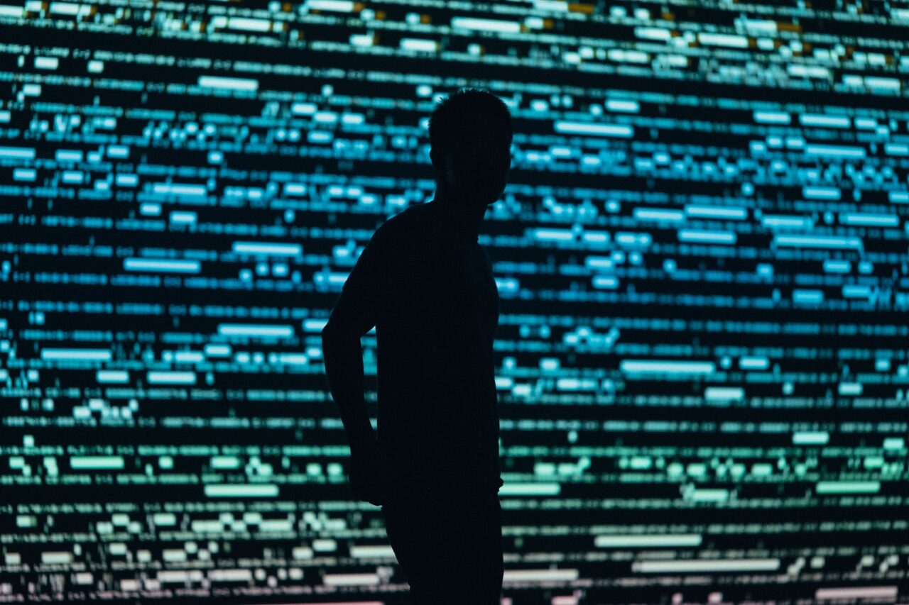 Sylwetka osoby stojącej na tle ekranu z kodem komputerowym.
