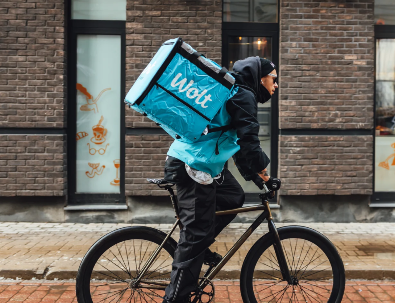 Kurier rowerowy z niebieskim, dużym plecakiem dostawczym jedzie przez miejską ulicę.