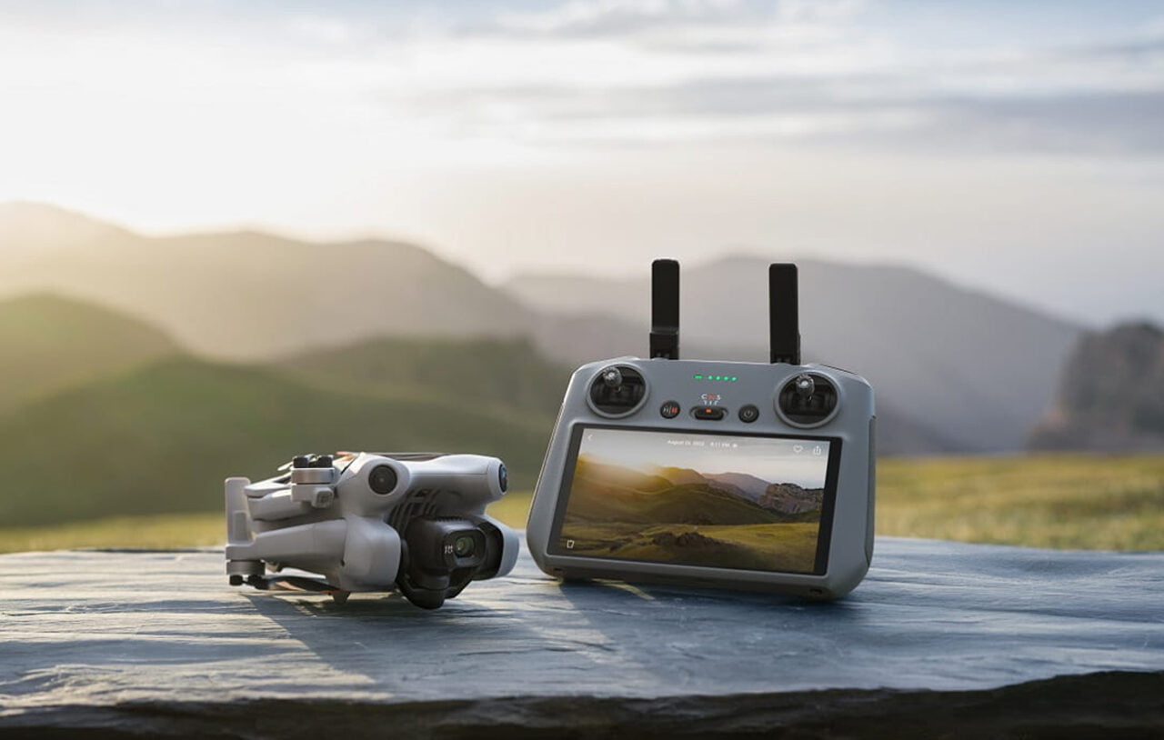 Dron DJI Mini 4 Pro Fly More Combo i kontroler zdalnego sterowania umieszczone na skałach na tle majestatycznych, zielonych wzgórz i wschodzącego słońca.