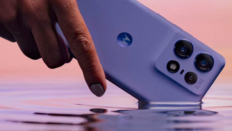 Ręka dotykająca tylną część fioletowego smartfona z logotypem Motoroli i trzema obiektywami aparatu, ukazana na tle odcieni różu.