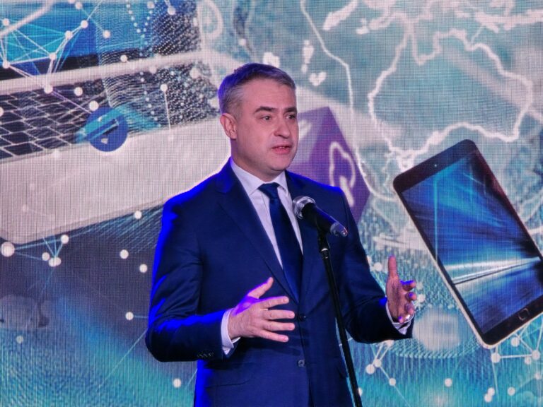 Minister cyfryzacji Krzysztof Gawkowski podczas 16. FG Time 2024. Mężczyzna w garniturze przemawiający przed mikrofonem z dużym ekranem z grafiką cyfrowych urządzeń i mapą świata w tle.