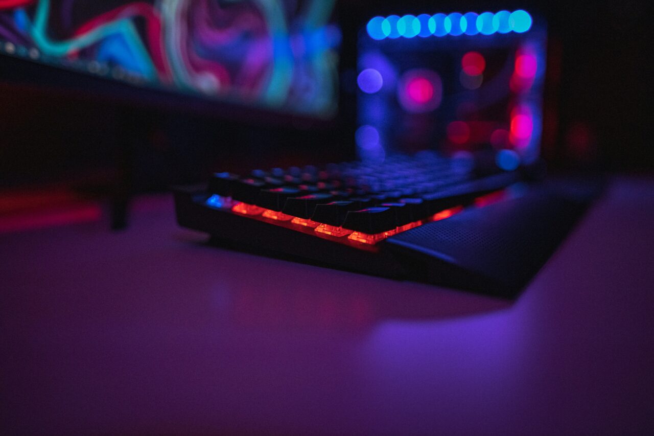 Podświetlana klawiatura gracza, którego zwolnienia w branży gier nie zainteresują. Komputerowa z czerwonymi światłami LED w ciemnym pokoju z rozmytym tłem przedstawiającym monitor i wydajny komputer z wewnętrznym oświetleniem.