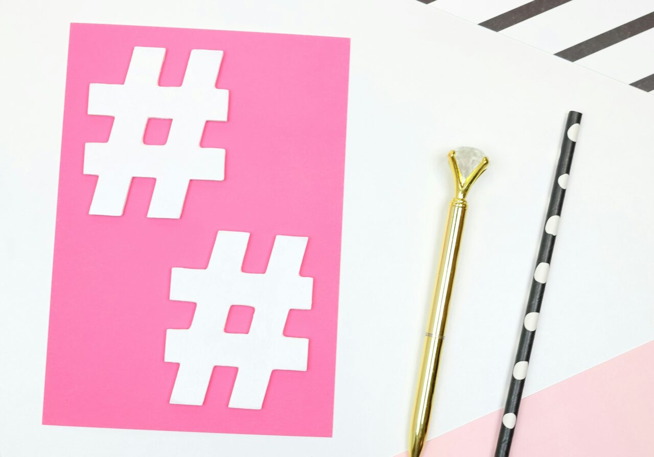 Um cartão rosa com recortes brancos do símbolo da hashtag, uma caneta dourada e um lápis preto com pontos brancos sobre fundo branco com elementos pretos e rosa.