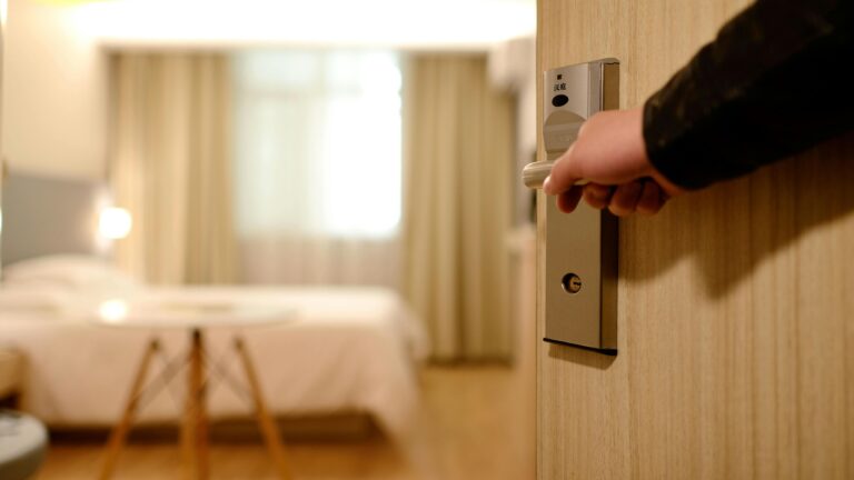 Ręka otwierająca metalowe drzwi z elektronicznym zamkiem do jasnego pokoju hotelowego.