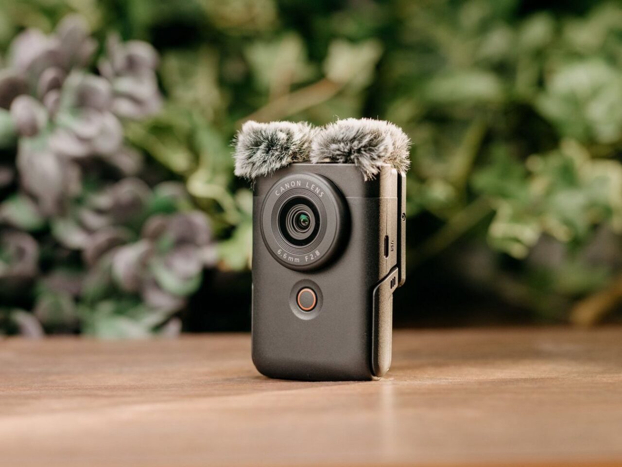 Aparat fotograficzny Canon z obiektywem i małym, puchowym mikrofonem zamocowanym na górze, stojący na drewnianym blacie z roślinnością w tle.