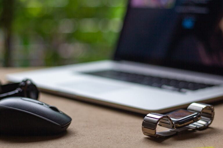 Biurko z rozmazanym w tle laptopem, na pierwszym planie ostra mysz komputerowa i rozmyty smartwatch na metalowej bransolecie.