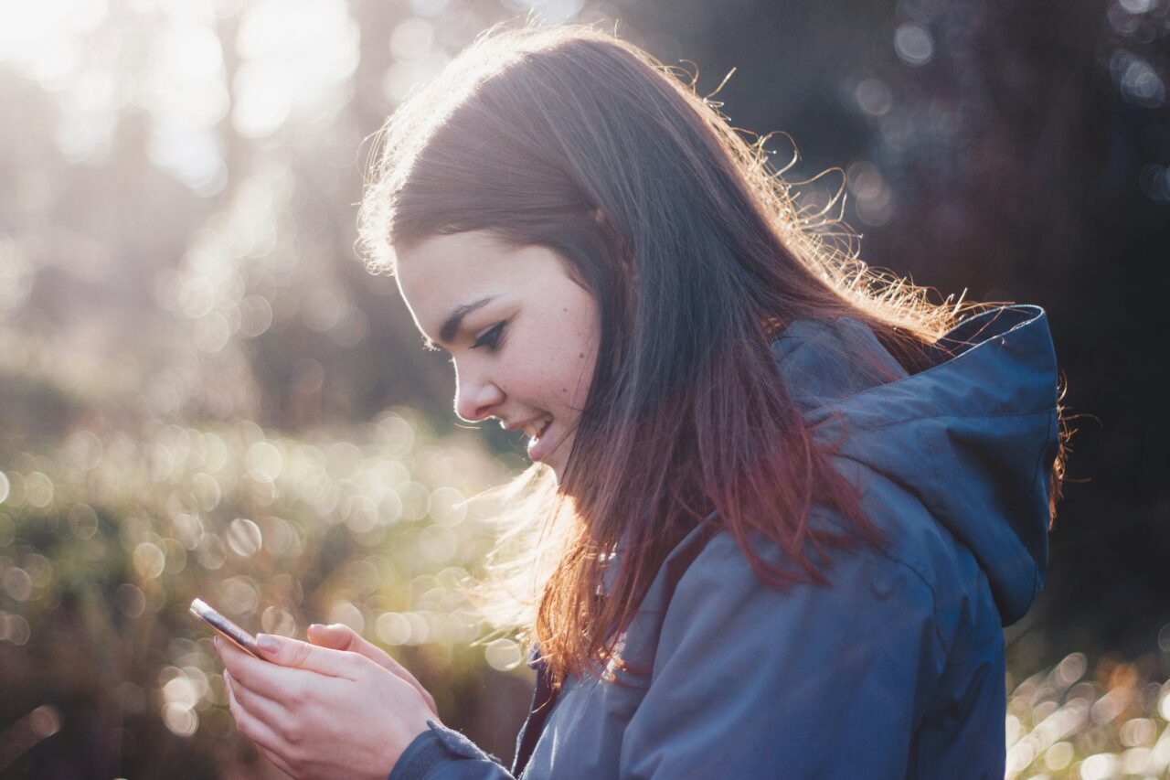 Młoda kobieta patrząca na smartfon, uśmiecha się, w tle rozmyte światło słoneczne.