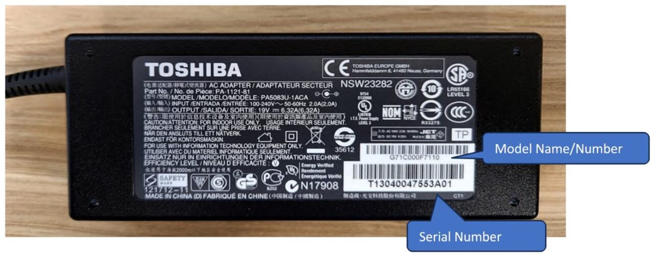 Czarny zasilacz Toshiba z etykietą zawierającą informacje o modelu i numerze seryjnym oraz różnymi certyfikatami i symbolami bezpieczeństwa, położony na drewnianej powierzchni.
