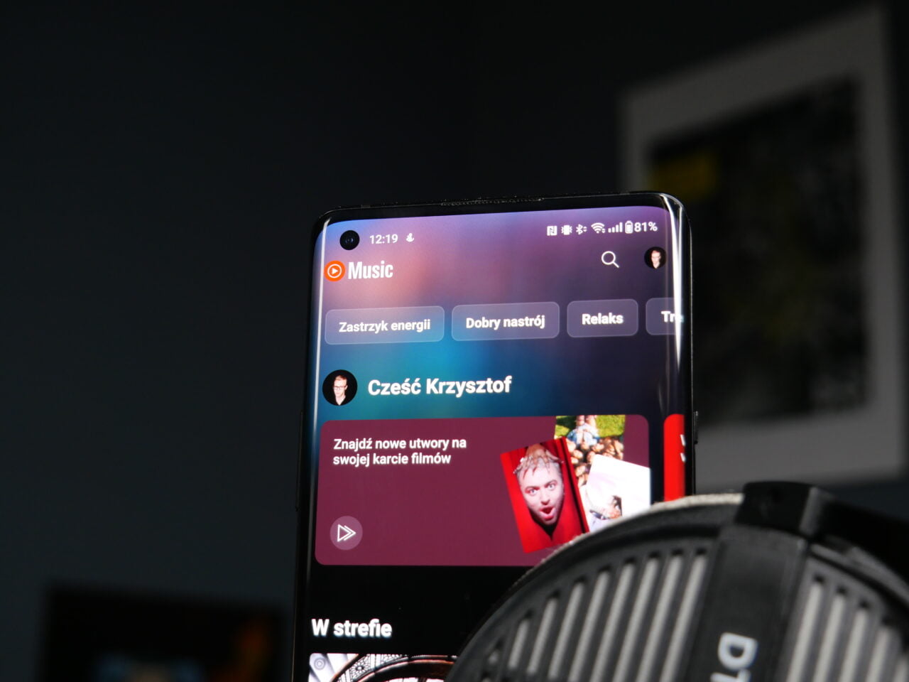Smartfon wyświetlający interfejs aplikacji muzycznej z grafiką użytkownika i opcjami wyboru playlisty, ustawiony na biurku przed nieostrym tłem.
