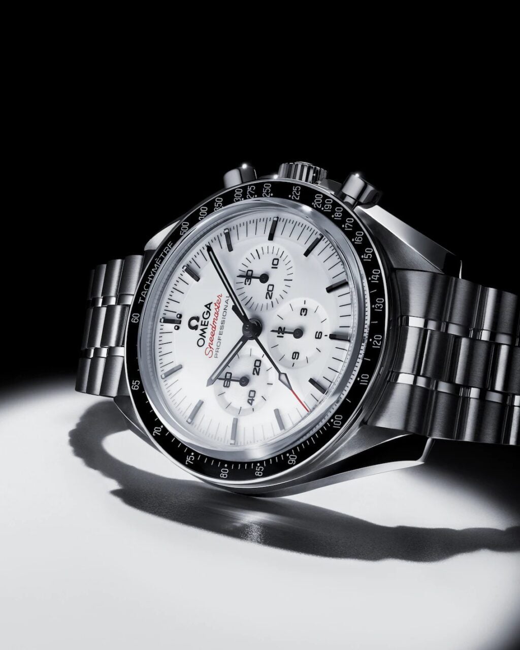 Zegarek męski Omega na metalowym pasku z białą tarczą, chronografem i czarnymi oznaczeniami, położony na białej powierzchni z cieniem.