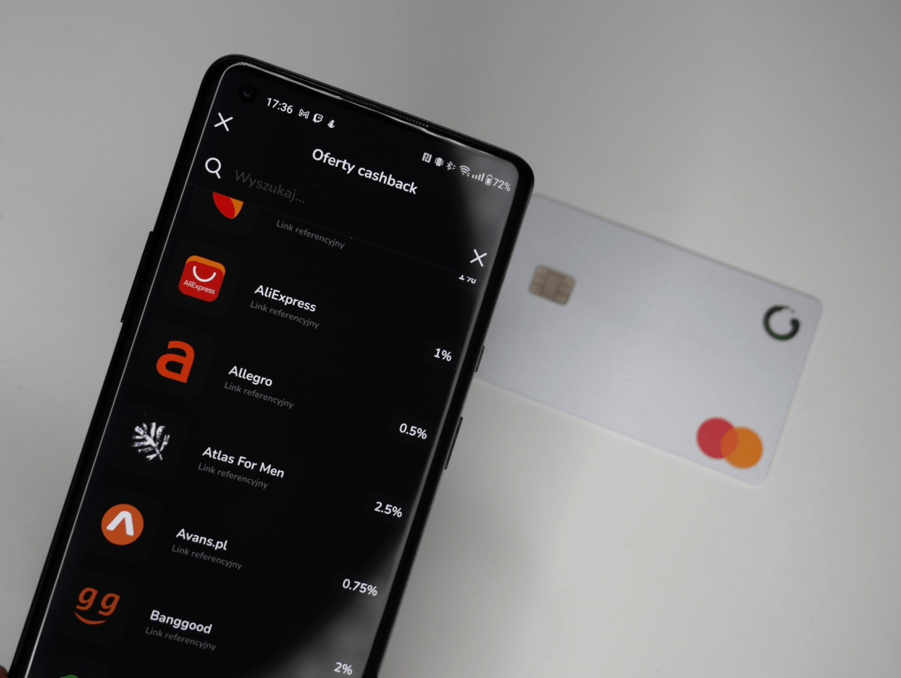 Smartfon wyświetlający aplikację ofert cashback z logotypami różnych sklepów internetowych, obok leży karta płatnicza.