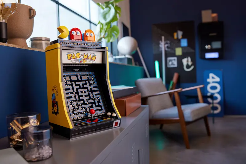 Maszyna do gier arcade Pac-Man w stylu retro postawiona na szafce, z pluszakami przedstawiającymi postacie z gry na górze, w nowoczesnym pokoju z ciemnoniebieskimi ścianami i stylowymi meblami.