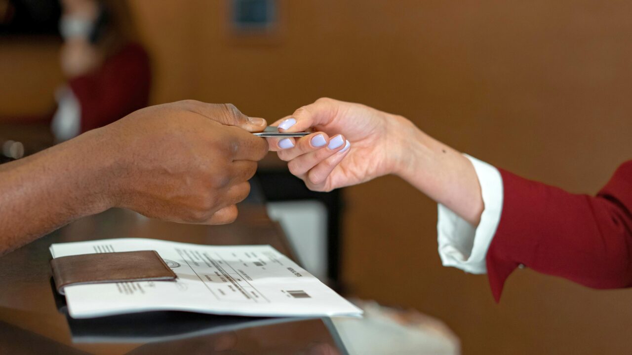 Dwie osoby wymieniają się kartą płatniczą przy hotelowej recepcji, na pierwszym planie widać papierową dokumentację i portfel.