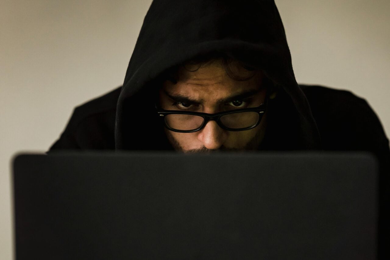 Haker. Mężczyzna w kapturze pracujący na laptopie, skupienie widoczne w jego oczach za okularami.