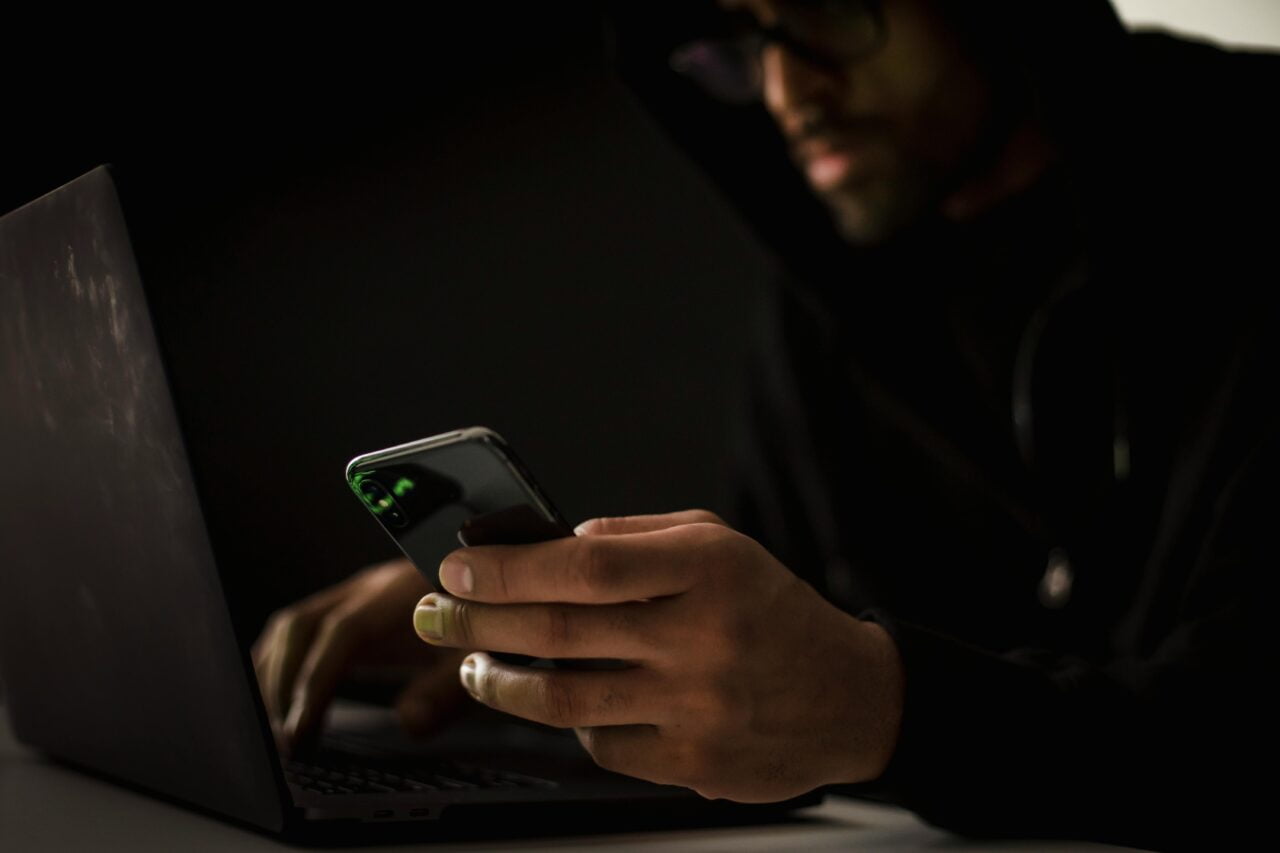 Haker. Mężczyzna w ciemnym ubraniu pracujący na laptopie i trzymający smartfon, z naciskiem na oświetlone dłonie, w tle ciemne otoczenie.