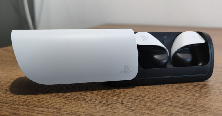 Biało-czarna stacja dokująca z otwartym pokrywem i widocznymi bezprzewodowymi słuchawkami PlayStation Pulse Explore, posiadającymi charakterystyczne oznaczenia firmy Sony, umieszczone na drewnianym blacie przy jasnej ścianie.