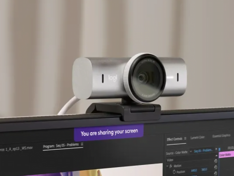 Uma webcam da Logitech colocada na borda superior de um monitor de computador com uma mensagem visível "Você está compartilhando sua tela" na tela do monitor.