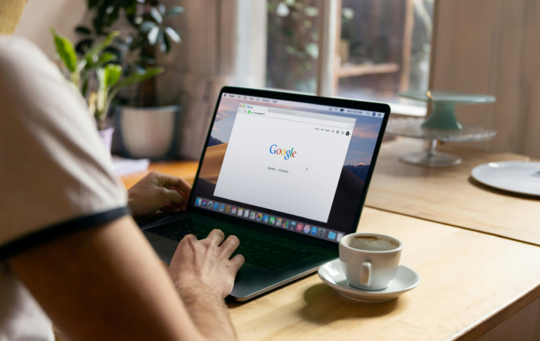 Osoba korzysta z laptopa z otwartą stroną główną Google, obok na biurku stoi filiżanka kawy.