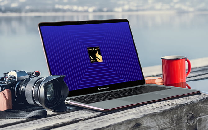 Um laptop em uma mesa de madeira em frente a um lago com uma tela aberta mostrando o logotipo do Snapdragon, ao lado de uma câmera e uma caneca vermelha.