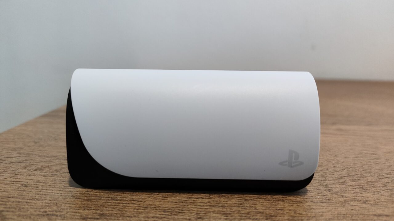 Biała stacja dokująca do  słuchawek PlayStation Pulse Explore z logo PlayStation, postawiona na drewnianym blacie przy jasnej ścianie.