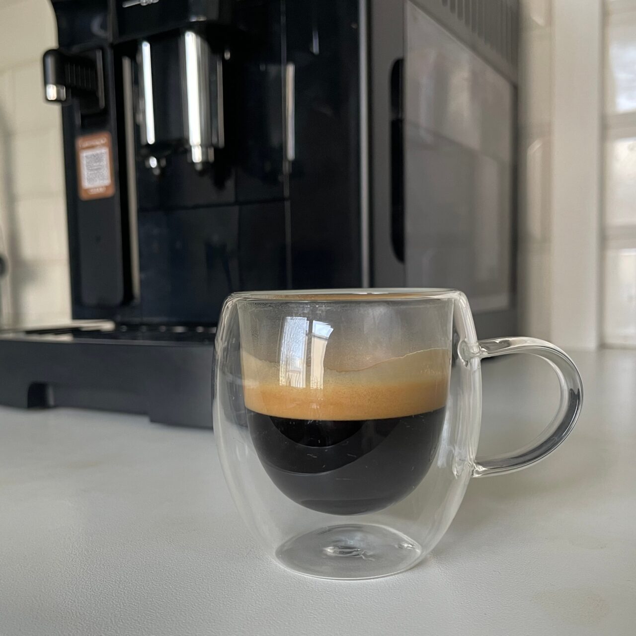 Filiżanka espresso na pierwszym planie z wyraźną cremą, w tle ekspres do kawy na kuchennym blacie.