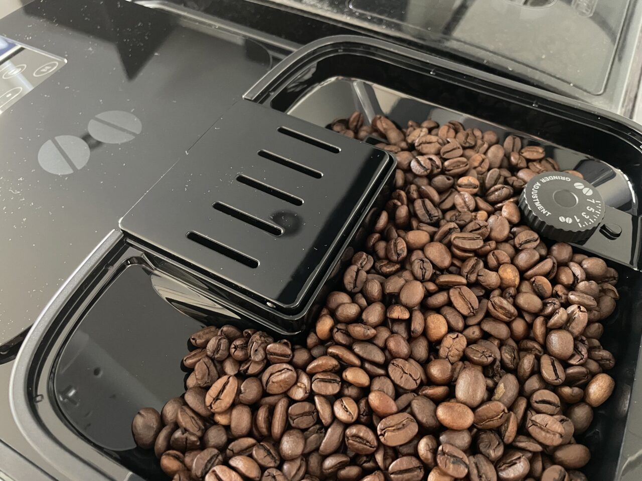Pojemnik na kawę z otwartą pokrywą wypełniony ziarnami kawy, umieszczony na czarnej, połyskującej powierzchni z fragmentami kuchenki indukcyjnej w tle.