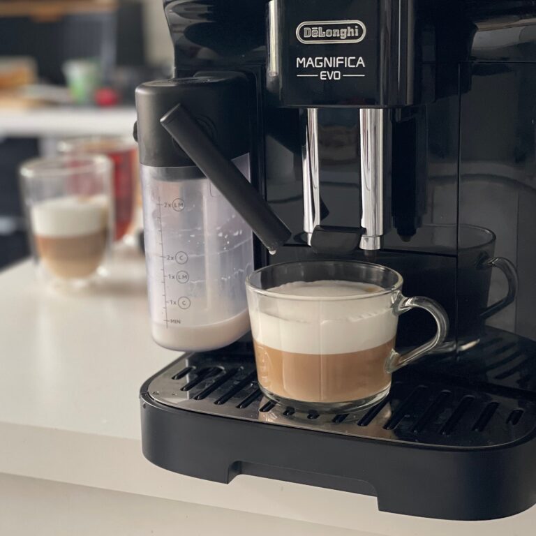 Ekspres do kawy marki De'Longhi model Magnifica EVO przygotowuje kawę latte w przeźroczystej filiżance. Obok stoi pojemnik z mlekiem do spieniania.