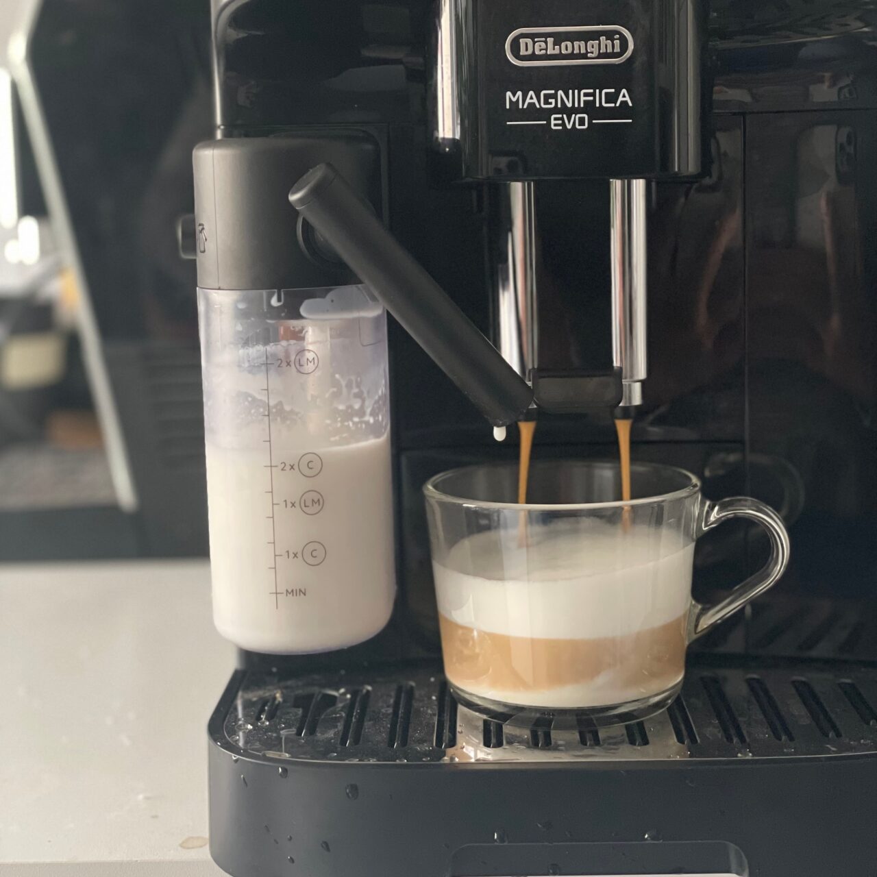 Ekspres do kawy De'Longhi Magnifica EVO zalewający espresso do przezroczystej filiżanki, obok pojemnik z mlekiem.