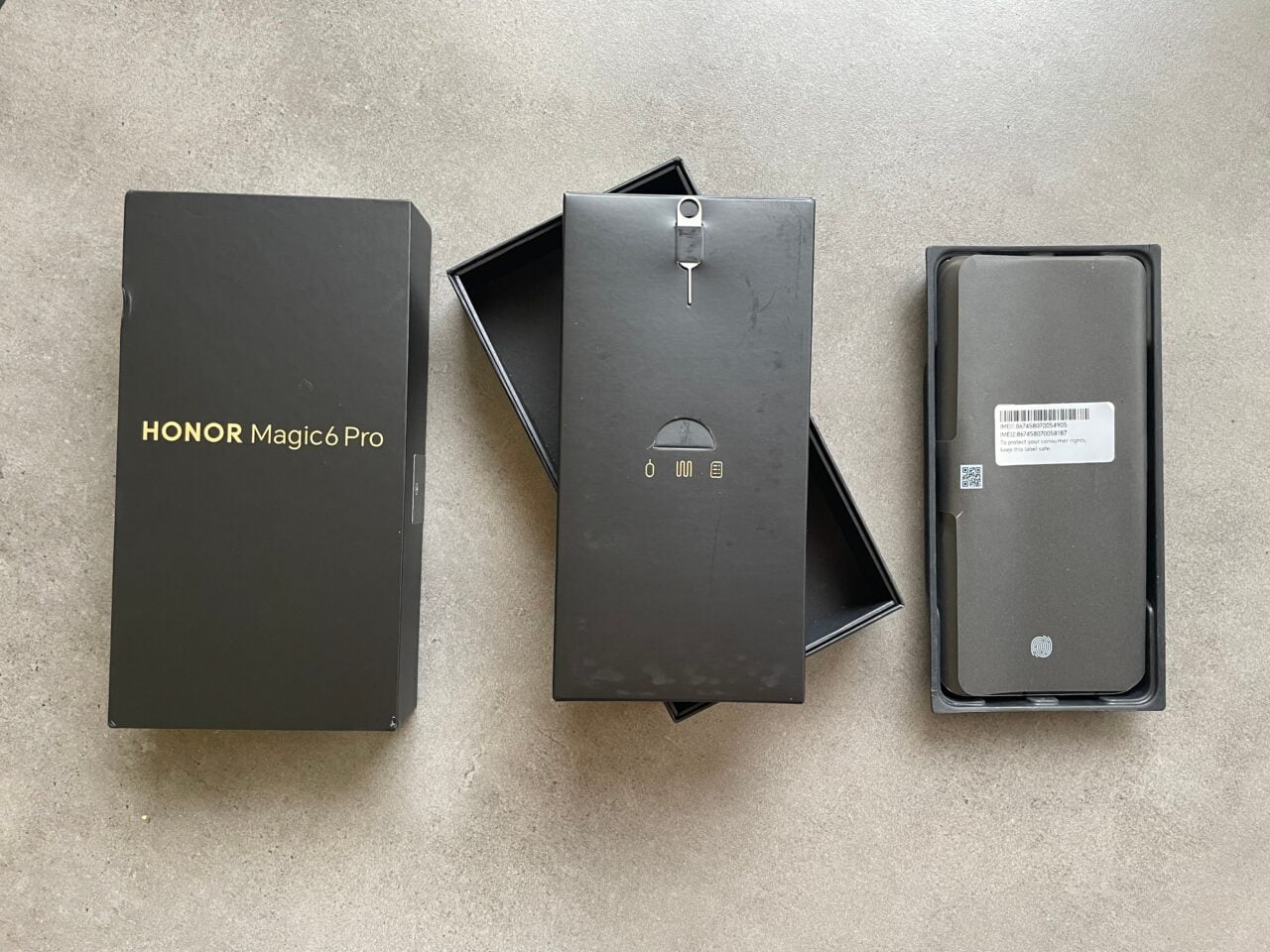 Zestaw pudełek telefonu komórkowego HONOR Magic6 Pro, w tym czarne pudełko zewnętrzne z napisem złotym, otwarte pudełko środkowe i wewnętrzne z plastikową tacą, na szarym tle.