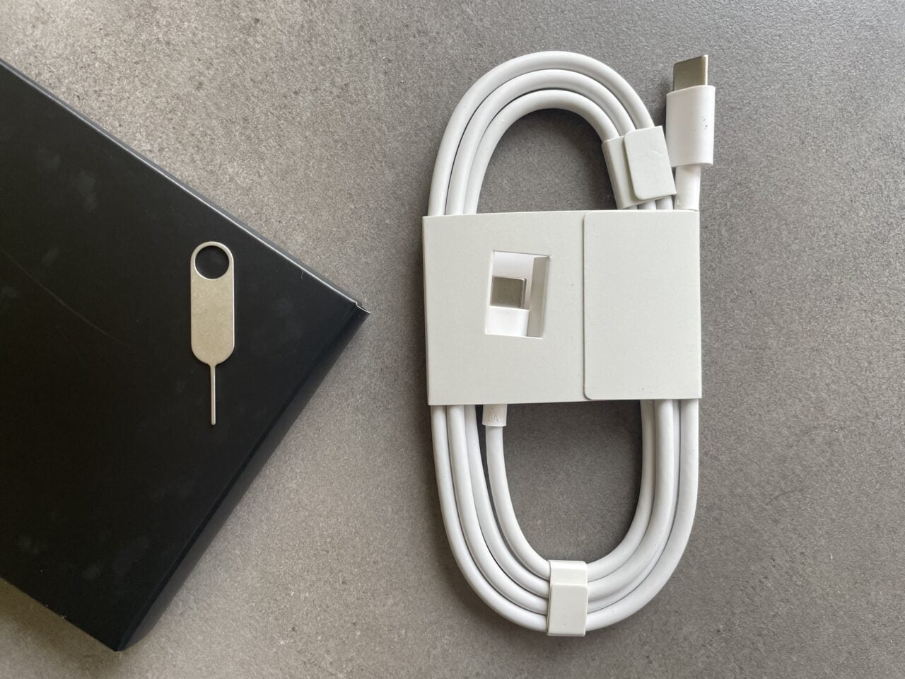 Biały przewód USB-C z organizatorem kabli i szpilką do wyciągania tacki SIM, obok czarne opakowanie na szarym tle.