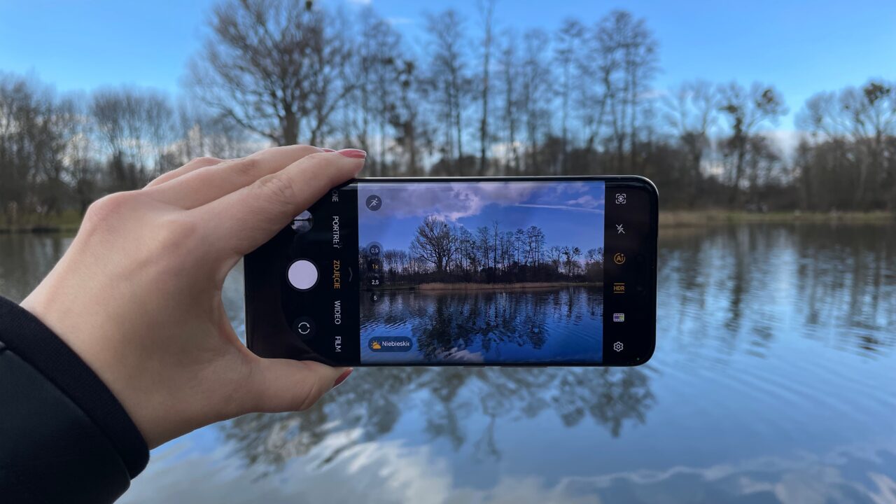 Osoba trzymająca smartfon, którym robi zdjęcie krajobrazu z odbiciem drzew i nieba w wodzie jeziora.