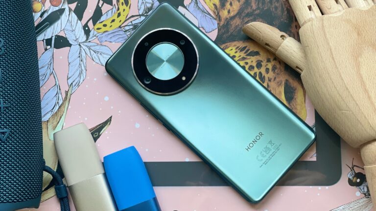 Smartfon Honor z dużym okrągłym modułem aparatu, leżący na kolorowej powierzchni obok głośnika, etui do słuchawek i drewnianej figurki.