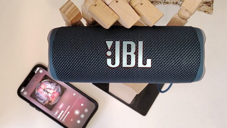 Głośnik przenośny JBL umieszczony na stole obok leżącego smartfona wyświetlającego ekran aplikacji muzycznej, z drewnianymi blokami w tle.