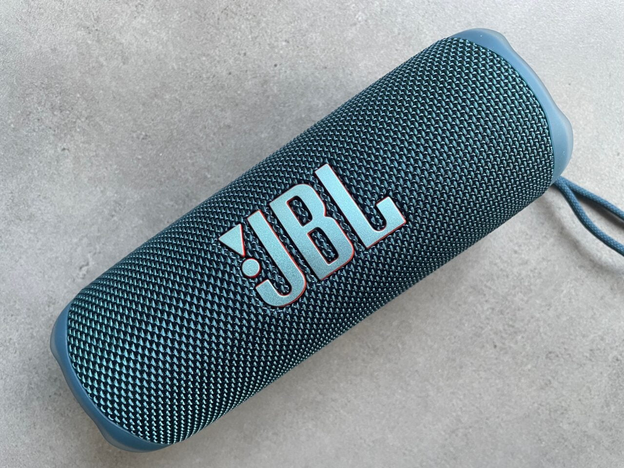 Przenośny głośnik Bluetooth marki JBL w kolorze turkusowym, leżący na jasnej powierzchni.