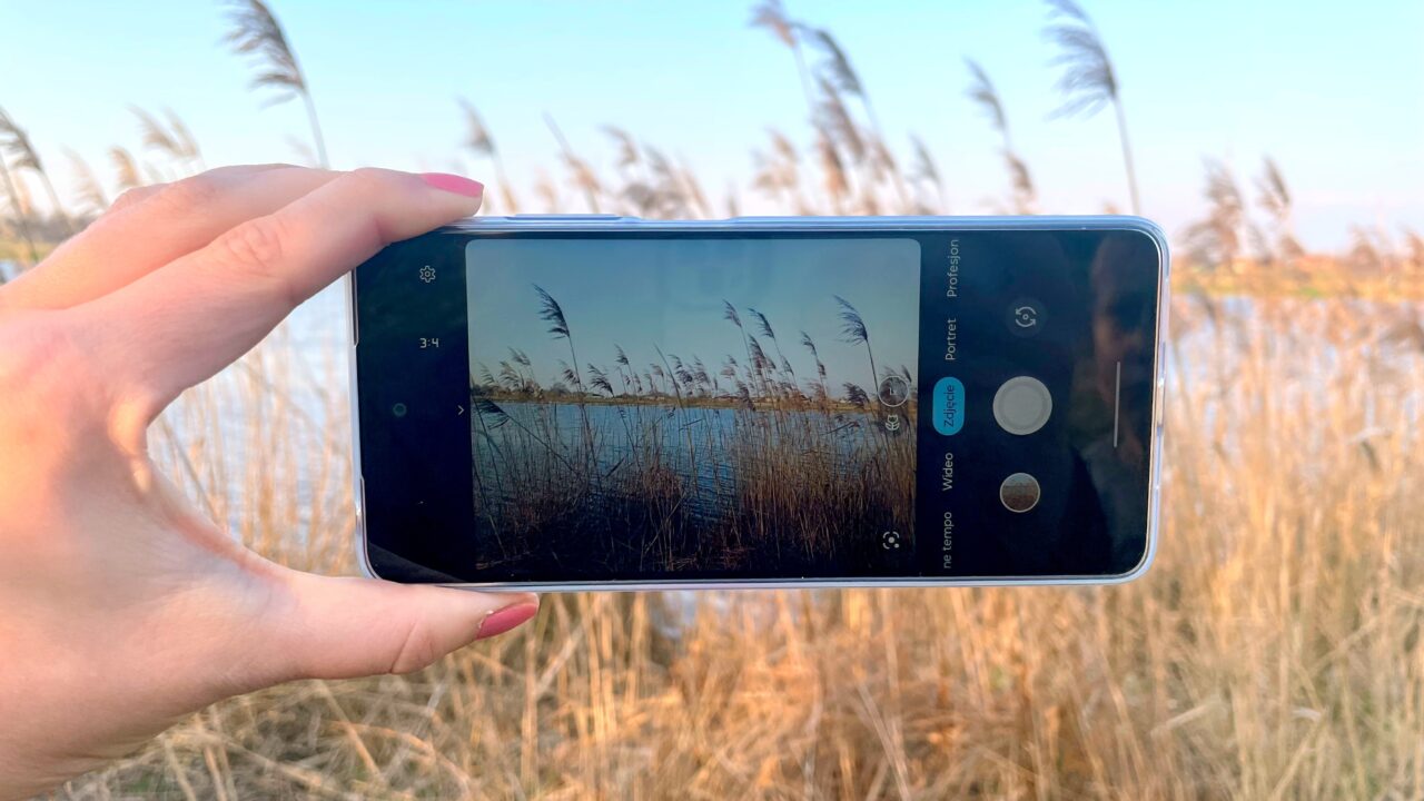 Ręka trzymająca smartfon, który fotografuje krajobraz z trzcinami nad jeziorem w świetle zachodzącego słońca.