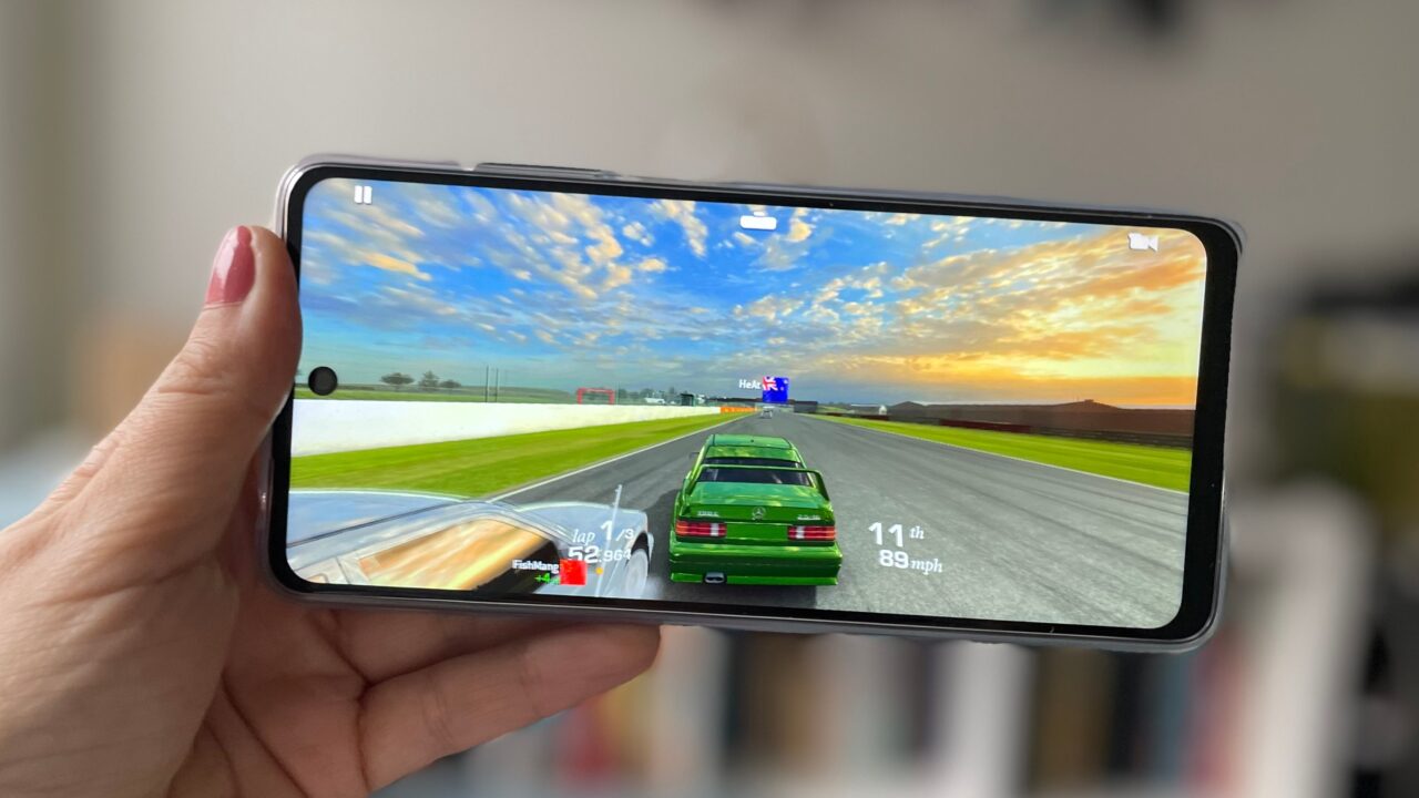 Osoba trzyma smartfon wyświetlający grę wyścigową z widokiem z pierwszej osoby, na ekranie zielony samochód wyścigowy na torze.