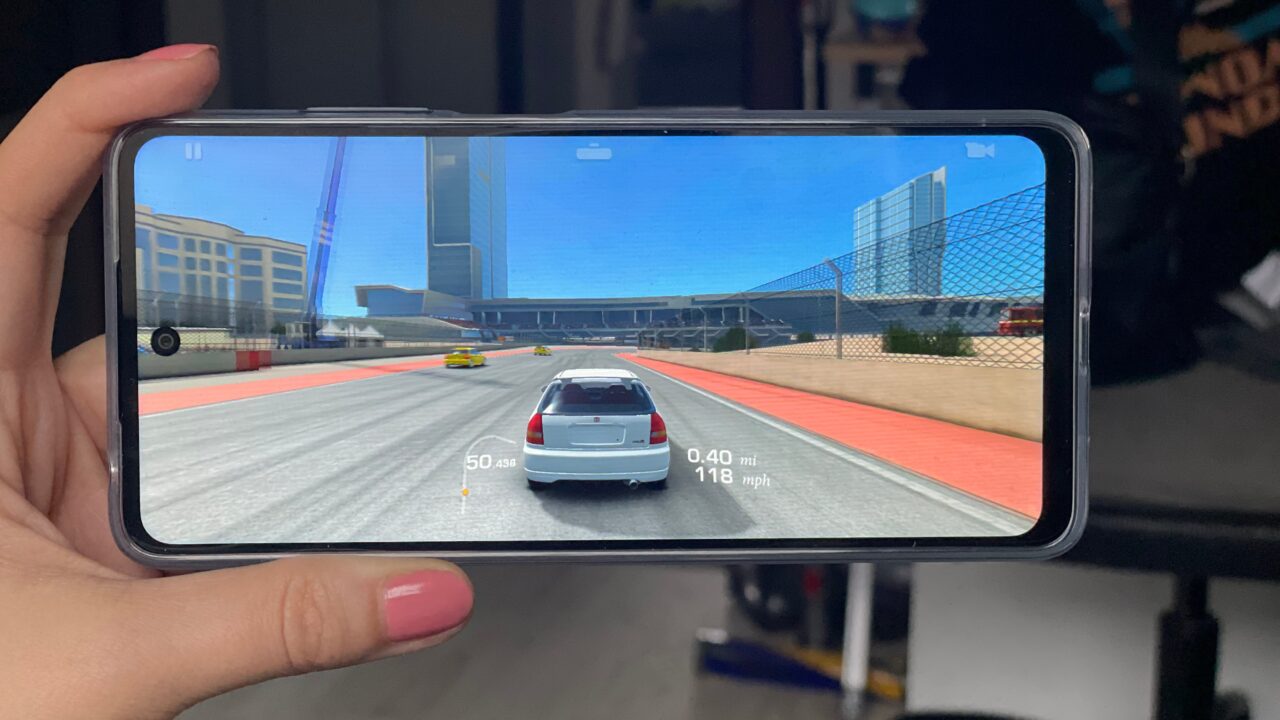Osoba trzyma smartfon wyświetlający grę wyścigową, na ekranie srebrny samochód jedzie po torze wyścigowym w mieście.