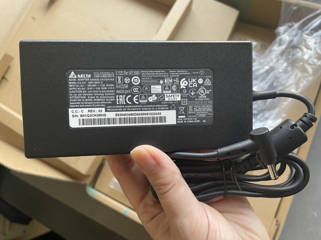 Czarny zasilacz sieciowy marki Delta Electronics trzymany w dłoni na tle kartonowych pudeł, z widocznymi etykietami i specyfikacjami na górnej stronie oraz złączem kabla zasilającego.