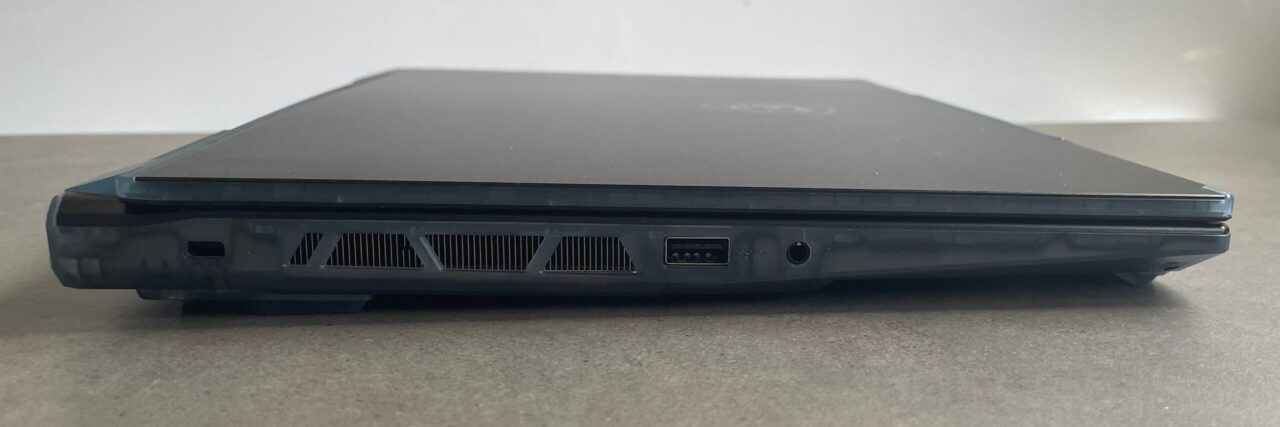 Zamknięty czarny laptop na szarym tle, widok od strony zawiasów z widocznymi portami USB, wentylacją i złączem audio.