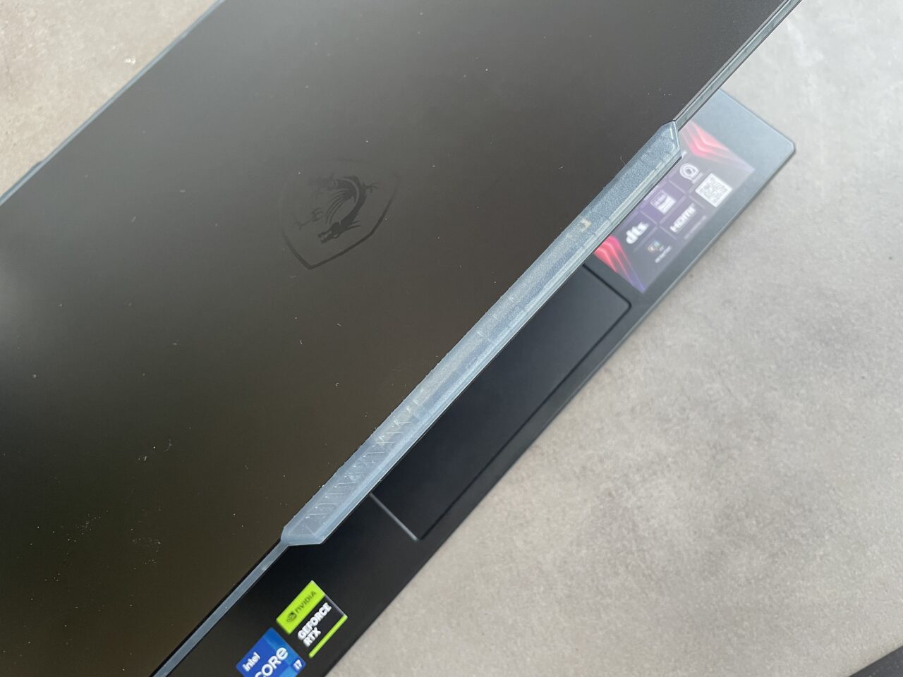 Zamknięty laptop gamingowy z ciemnym wykończeniem i logiem w kształcie smoka na pokrywie, z naklejkami informacyjnymi o specyfikacji na dolnej krawędzi ekranu.