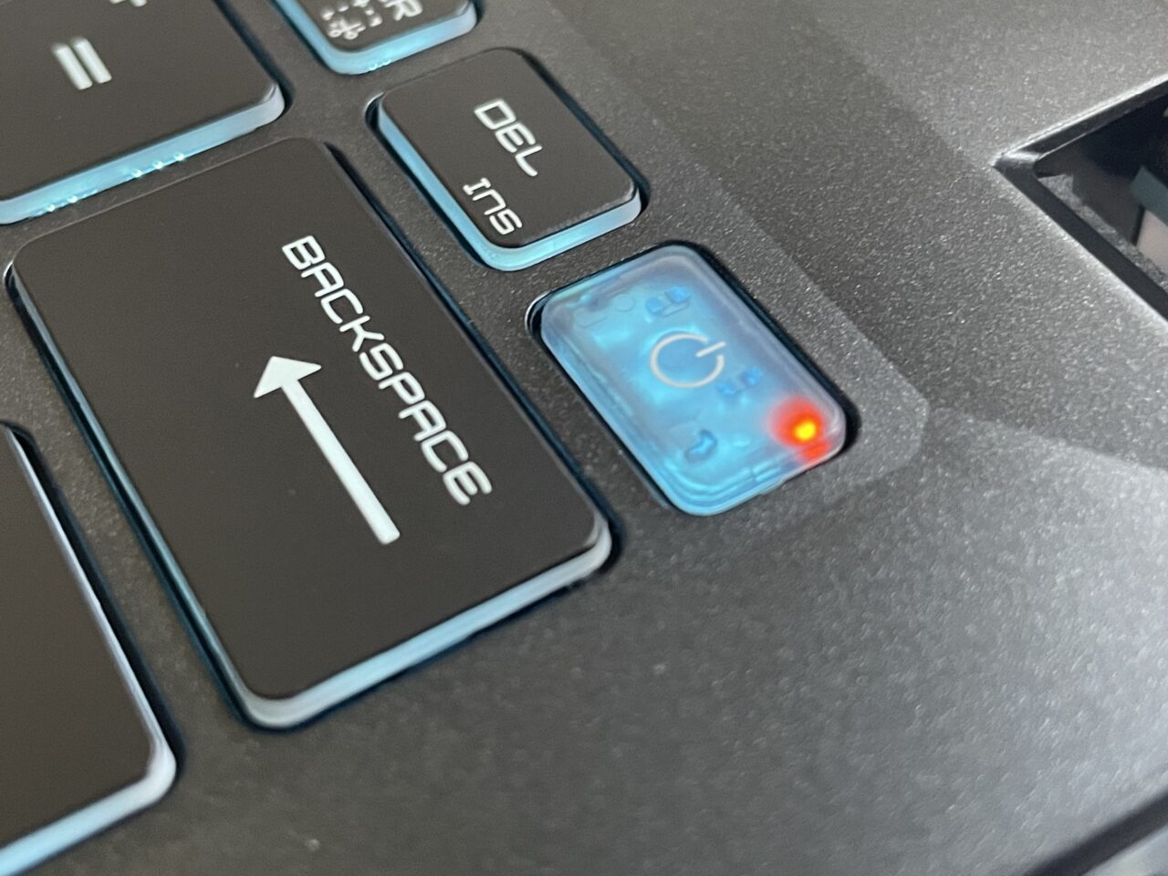Część klawiatury laptopa z wyróżnionym przyciskiem 'Backspace' i świecącym czerwonym światłem wskaźnika zasilania.