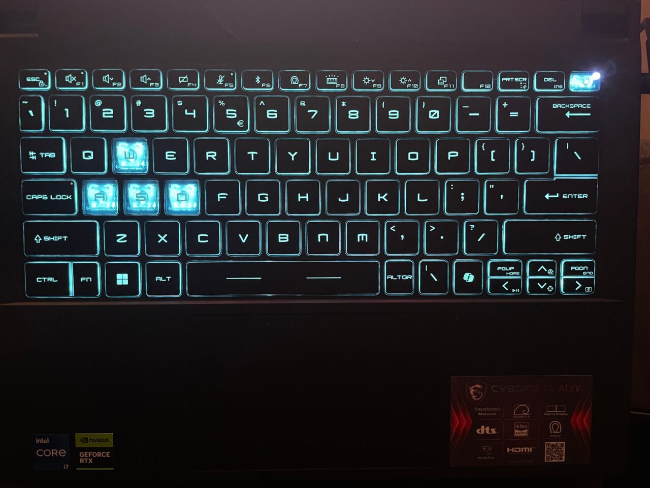 Podświetlana klawiatura komputerowa z wyeksponowanymi klawiszami WASD i znakami graficznymi na klawiszach funkcji, oraz naklejkami Intel Core i7 i NVIDIA GeForce RTX na dolnej ramce.