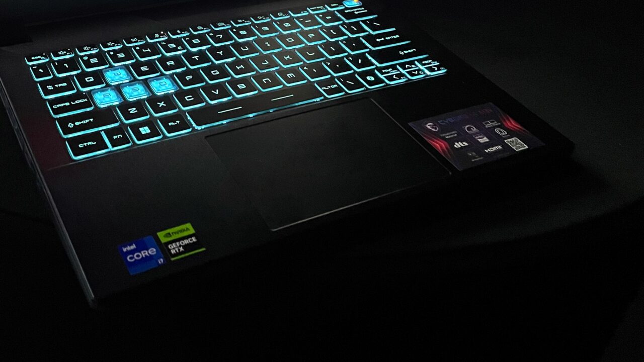 Laptop z podświetlaną klawiaturą na niebiesko w ciemnym otoczeniu, z naklejkami Intel Core i7 i NVIDIA GeForce RTX.