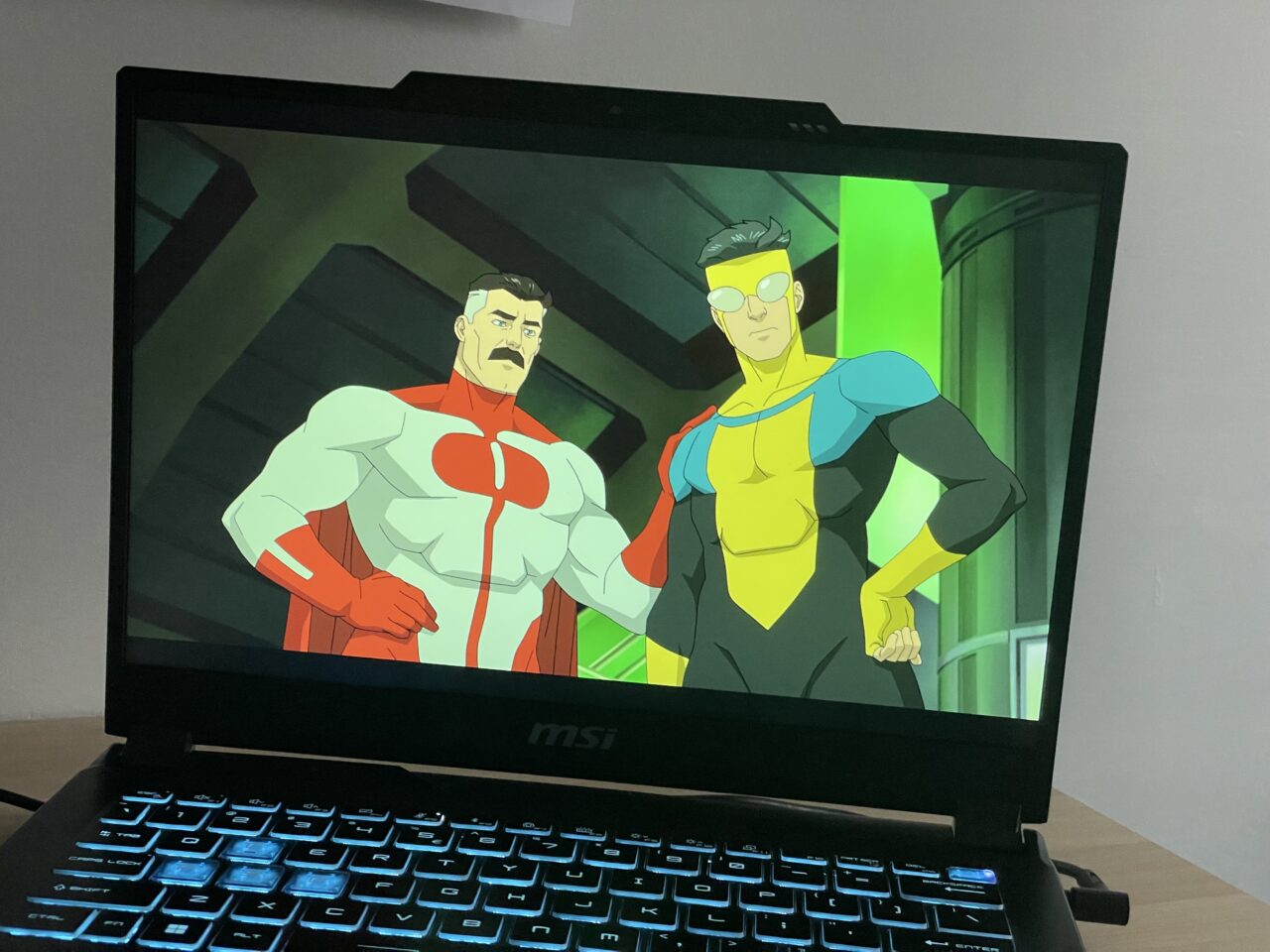 Laptop MSI wyświetlający kreskówkę z dwoma męskimi postaciami superbohaterów, jeden w czerwono-białym, drugi w żółto-niebieskim stroju, na tle zielonej poświaty.